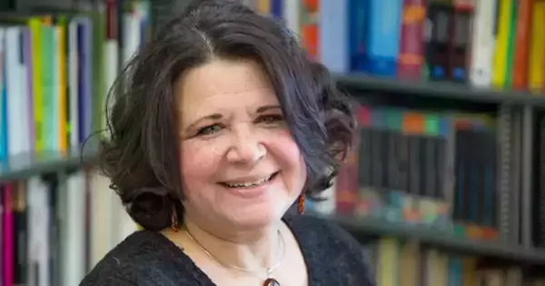 A woman with shoulder-length, dark curly hair smiles at the camera. She wears a dark shirt and a pendant necklace. Bookshelves filled with colorful books are visible in the background.