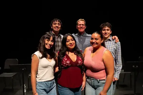 Six smiling people: three women in white, maroon, and pink tops; three men in plaid and blue shirts, on a dark stage.