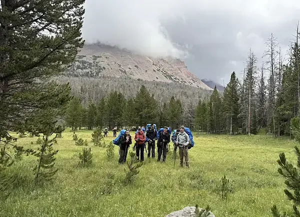 LSSP 10 WONDER: Group of hikers with blue backpacks stand in a grassy field with a mountain in the background.
